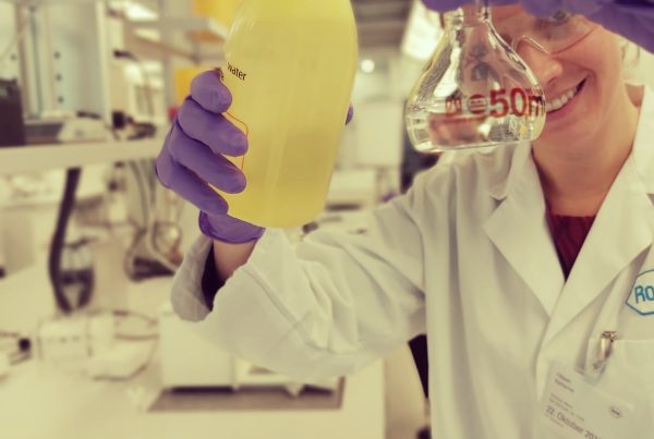 Eine blonde Person trägt einen weißen Kittel und lila Handschuhe. Sie hält eine Flasche und ein Reagenzglas in das Bild. Im Hintergrund ist ein Labor zu sehen.
