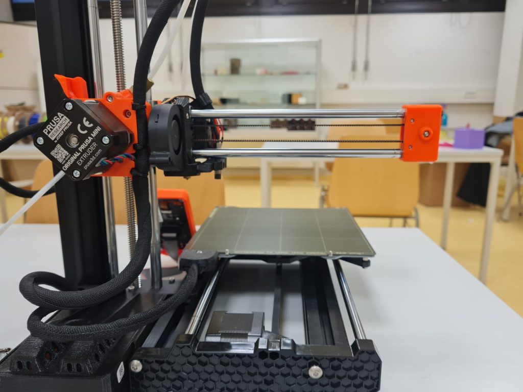 3D Drucker in schwarz und neon orange steht auf einem weißen Tisch in einem Klassenzimmer. Der Druker hat eine Blatte mit aufgezeichneten quadratischen weißen Linien. Auf der Linken Seite sieht man eine stehende Stange und davon ab geht horizontal eine Stange weg die orangene Elemente hat und in der Mitte aus Metall ist. Die Horizontale Stange is mit der vertikalen Stange und der Platte mit 2 Kabeln verbunden. Die Platte ist auf 2 metallenen Schienen befestigt und ist im Foto ganz nach vorne ausgerchtet. Die Oberfläche an der Hinterseite des Druckers ist wie Bienenwaben 6 eckig geformt und aus schwarzem Plastik.
