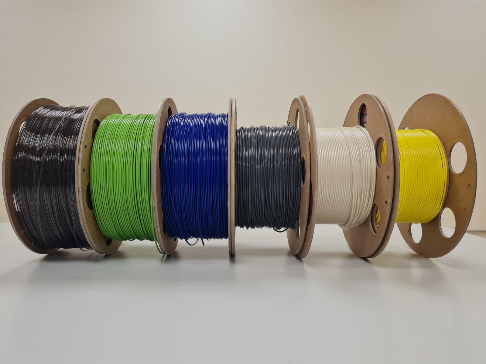 6 Filament Rollen mit verschiedenen Farben 3D-Druckfilament: schwarz, grün, blau, grau, weiß und gelb. Sie sind dicht nebeneinander auf einem weißen Tisch vor einem weißen Hintergrund platziert.
