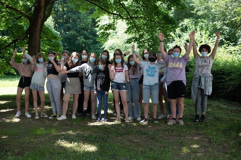 Eine Gruppe von Personen steht auf grünem Gras vor mehreren grünen Bäumen und einem braunen Baumstamm. Die 18 Personen heben ihre Arme hoch, tragen alle Mund-Nasenmasket und tragen verschiedenfarbene Kleidung.