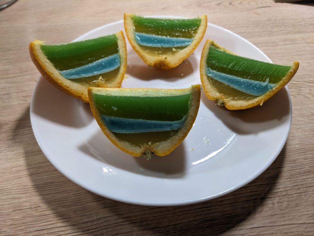 4 Orangenschnitze auf einem weißen Teller. Die Orangen haben grün, braun, blau, braune Streifen.
