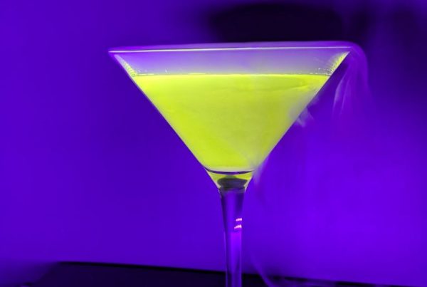 Ein Cocktailglas mit dampfenden gelbneon leuchtendem Inhalt vor lila abgedunkeltem Hintergrund.