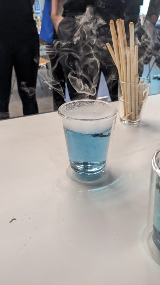 Auf einem Tisch steht ein Glas mit blauer durchsichtiger Flüssigkeit. Die Flüssigkeit dampft und hat Schaum auf der Overfläche. Im Hintergrund stehen Grissinis in einem Glas. Dahinter stehen 3 Menschen mit schwarzen Hosen.