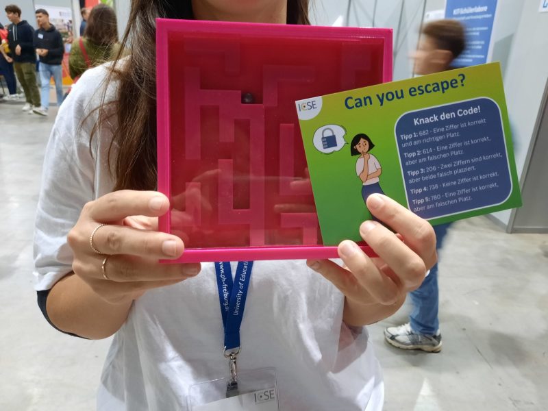 Eine Person hält ein 3D-gedrucktes pinkes Labyrinth mit kleiner Kugel in die Kamera. Dazu einen Flyer auf dem steht "Can you escape? Knack den Code!"