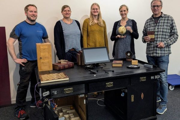 Eine Gruppe von 5 Personen steht um einen großen schwarzen Schreibtisch herum. Auf dem Schreibtisch befinden sich Gegenstönde, wie ein Schachbrett, eBücher, eine Uhr und ein Bildschirm.