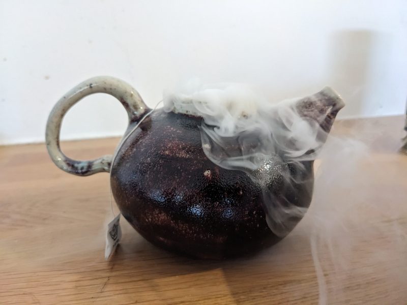 Eine Teekanne steht auf einem Holztisch. Aus ihr kommt Rauch.