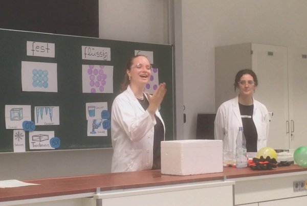 Eine Person in weißem Kittel, mit Schutzbrille und blondenlangen Haaren steht vor einer Schultafel und gebärdet das Wort "Danke".