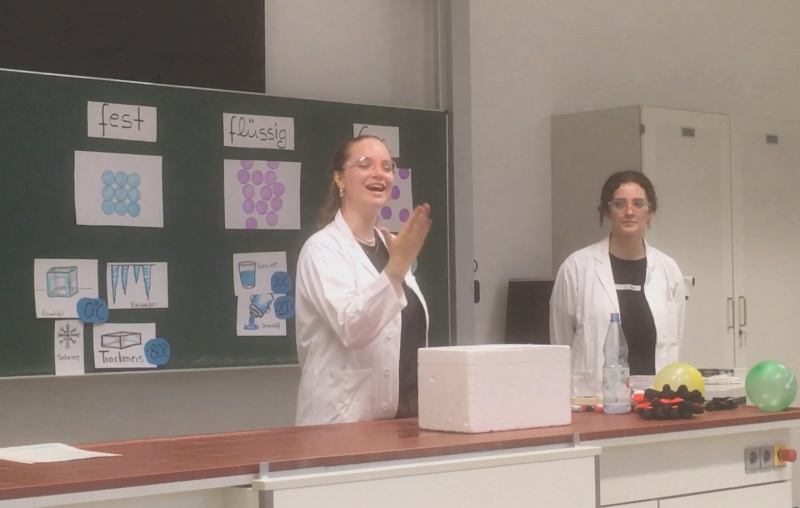 Eine Person in weißem Kittel, mit Schutzbrille und blondenlangen Haaren steht vor einer Schultafel und gebärdet das Wort "Danke".