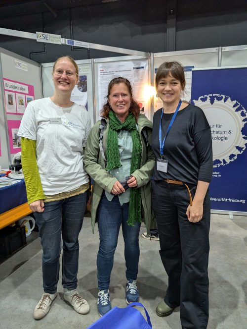 Das ICSE Science Factory Team steht vor dem Stand: 3 Personen, eine mit blonden Haaren, und 2 mit braunen Haaren stehen vor einer Ecke in der viele Poster hängen.