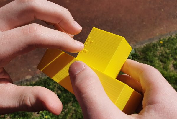 Ein senf-gelbes Objekt, bestehend aus zwei gelben Rechtecken (das Untere größer als das Obere) werden von zwei Händen gehalten. Der Zeigefinger der linken Hand fährt über Braille, die auf die linke Seite des oberen Rechtecks gedruckt ist. Im Hintergrund ist Gras zu sehen und eine braune Wand.