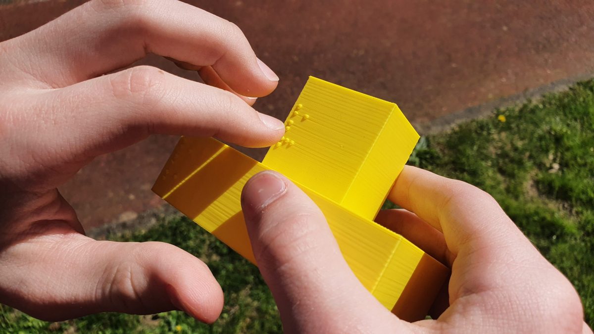 Ein senf-gelbes Objekt, bestehend aus zwei gelben Rechtecken (das Untere größer als das Obere) werden von zwei Händen gehalten. Der Zeigefinger der linken Hand fährt über Braille, die auf die linke Seite des oberen Rechtecks gedruckt ist. Im Hintergrund ist Gras zu sehen und eine braune Wand.