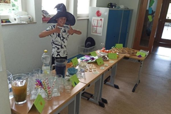 Ein Jugendlicher steht hinter einem Tisch voller Essen und Getränke, trägt einen schwarzen Hexenhut und reckt die Daumen in die Höhe.