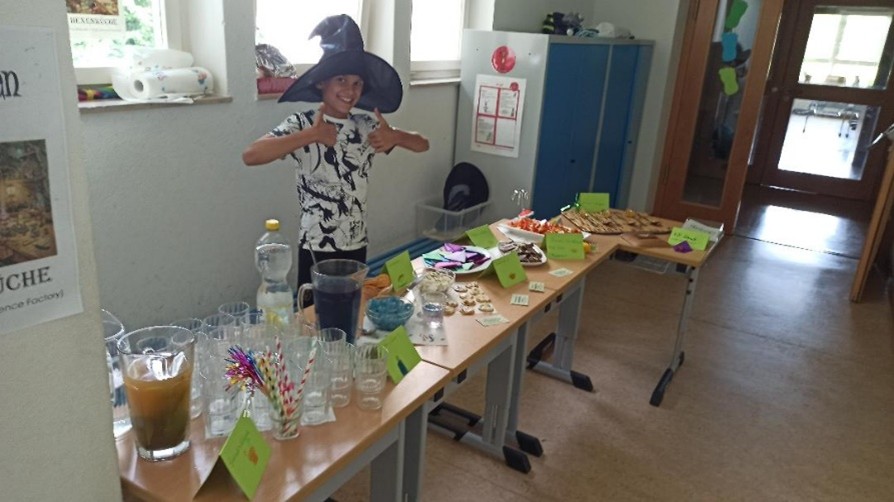 Ein Jugendlicher steht hinter einem Tisch voller Essen und Getränke, trägt einen schwarzen Hexenhut und reckt die Daumen in die Höhe.