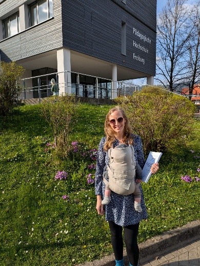 Eine blonde Person mit Manduka und Baby auf der Brust steht vor einem grauen Gebäude und hält einen gebundenen Stapel Papier in der Hand. Sie trägt eine Sonnenbrille und lächelt. Auf dem Gebäude steht Pädagogische Hochschule Freiburg.