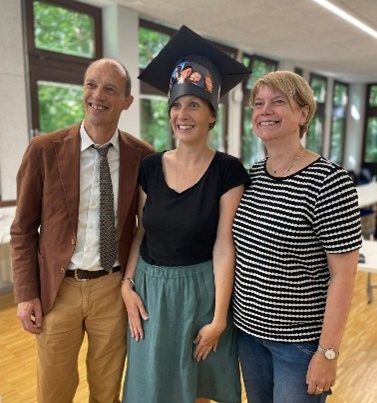 Katharina steht zwischen 2 weiteren Personen: einem Menschen mit braunem Sakko und Krawatte auf der linke Seite und einer Person mit blonden kurzen Haaren und einem gestreiften T-Shirt auf der rechten Seite. Katharina trägt einen großen schwarzen Hut mit Fotos darauf. Alle lächeln in die Kamera.