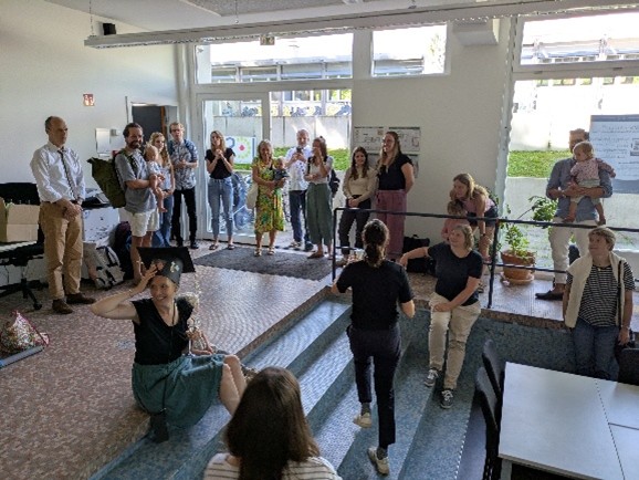 In einem Büro sind viele Menschen versammelt. In der Mitte sitzt Katharina mit einem schwarzen großen hut mit Bildern darauf. Neben ihr sitzt ein Kind.