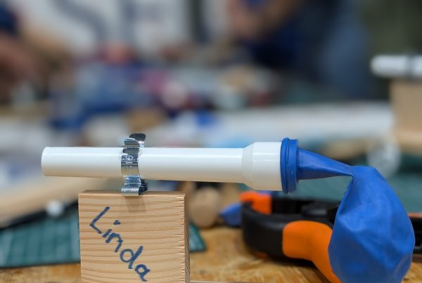 Ein Fahrzeug mit 4 weißen Rädern, einem Holzrumpf und einem blauen Luftballon, der über ein weißes Plastikrohr gestülpt ist steht auf einem Holztisch. Im Hintergrund sind mehrere Bastelstationen zu sehen, auf denen die Materialien und Werkzeuge für weitere Fahrzeuge stehen.