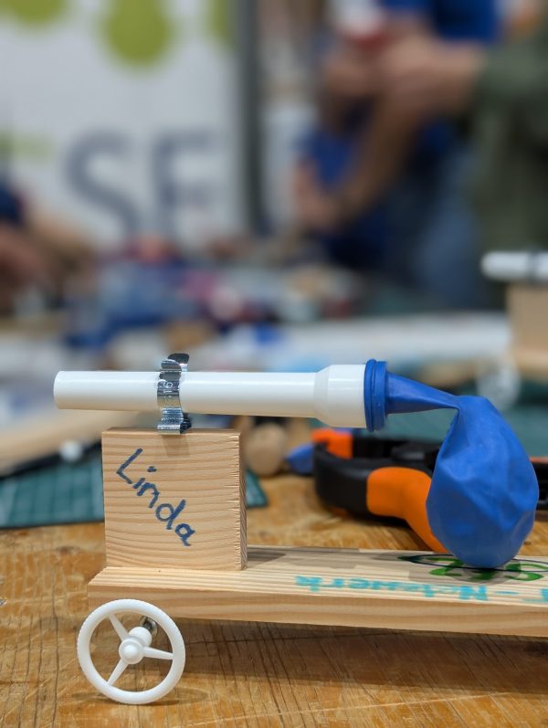 Ein Fahrzeug mit 4 weißen Rädern, einem Holzrumpf und einem blauen Luftballon, der über ein weißes Plastikrohr gestülpt ist steht auf einem Holztisch. Im Hintergrund sind mehrere Bastelstationen zu sehen, auf denen die Materialien und Werkzeuge für weitere Fahrzeuge stehen.