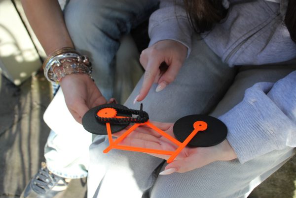 Ein knall orangenes Fahrradmodell mit schwarzen Rädern liegt in der Hand von einer Person, die mit dem Finger darüber fährt. Eine zweite Person sitzt neben der Person.