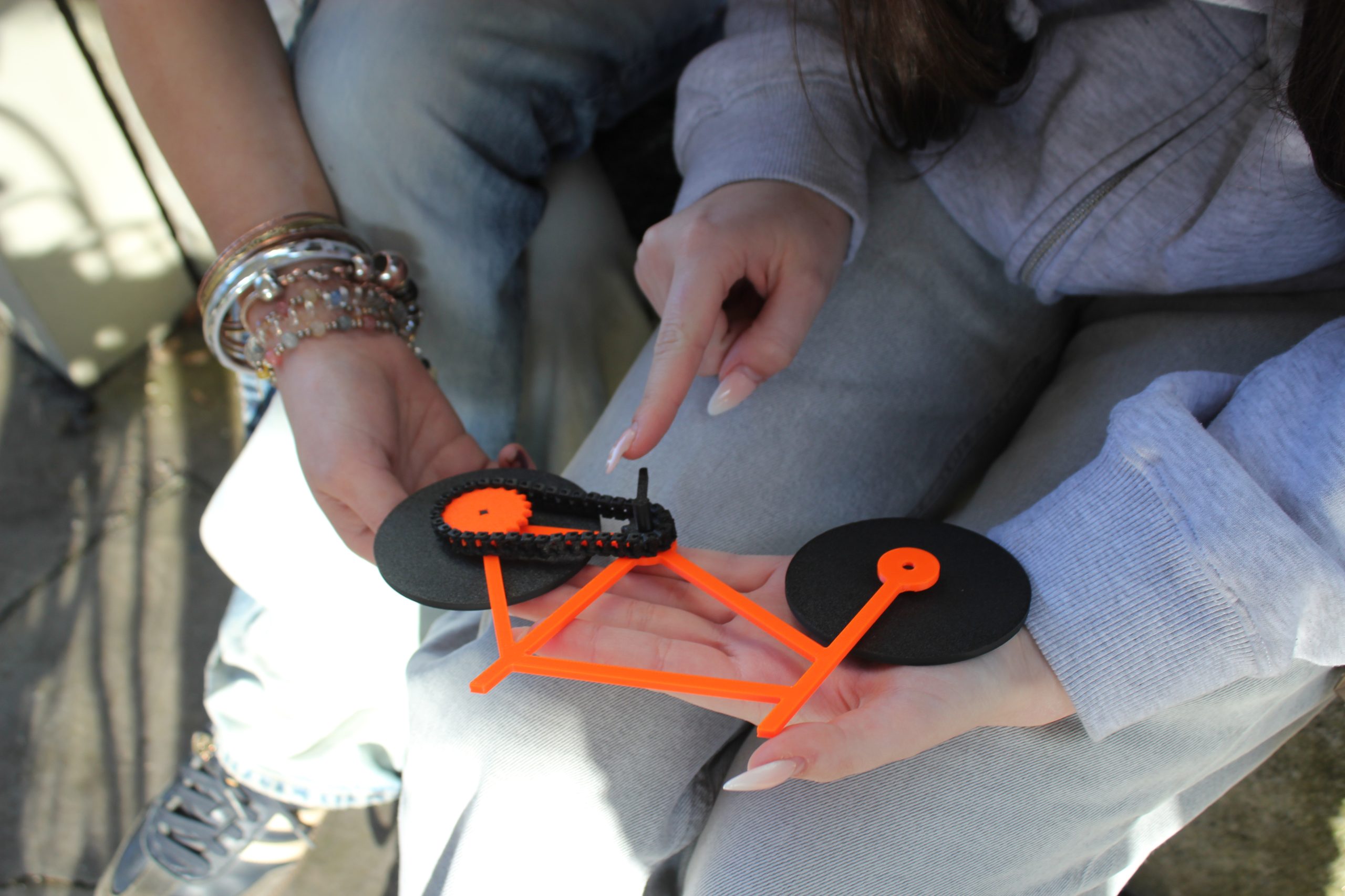 Ein knall orangenes Fahrradmodell mit schwarzen Rädern liegt in der Hand von einer Person, die mit dem Finger darüber fährt. Eine zweite Person sitzt neben der Person.